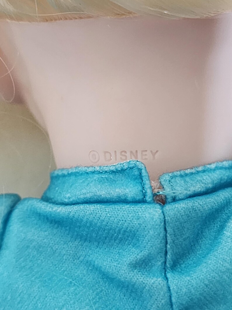 Puede incluir: Primer plano del cuello y los hombros de una mu&ntilde;eca. La mu&ntilde;eca tiene el pelo rubio y lleva un vestido turquesa. La palabra "&copy; DISNEY" est&aacute; impresa en la parte posterior del cuello de la mu&ntilde;eca.