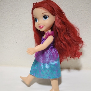 Puede incluir: Mu&ntilde;eca de un personaje con cabello rojo, ojos azules y un vestido morado y turquesa. La mu&ntilde;eca est&aacute; de pie con un pie levantado, como si caminara. El vestido tiene una falda turquesa con un dise&ntilde;o de remolino.