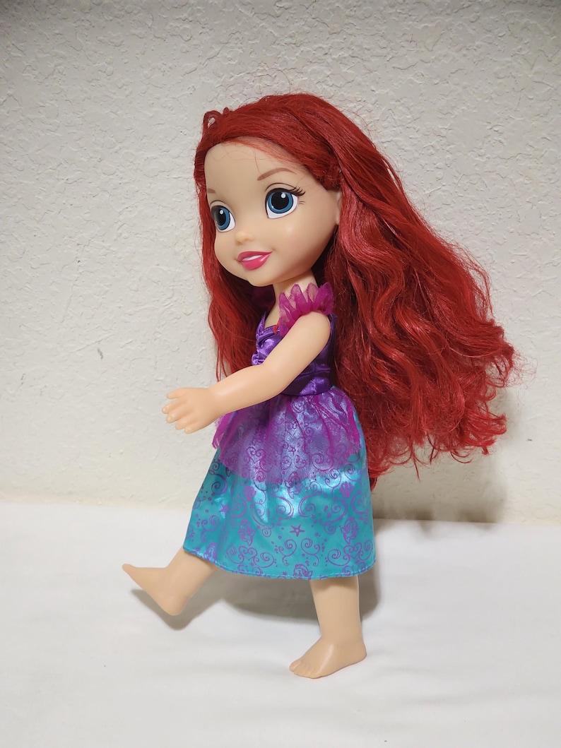 Puede incluir: Mu&ntilde;eca de un personaje con cabello rojo, ojos azules y un vestido morado y turquesa. La mu&ntilde;eca est&aacute; de pie con un pie levantado, como si caminara. El vestido tiene una falda turquesa con un dise&ntilde;o de remolino.