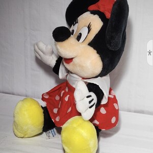 May include: A plush Minnie Mouse doll, featuring black fur, a red polka-dot skirt, and a matching bow. The doll has large yellow feet, white gloves, and a white collar. The doll is sitting and appears to be waving.