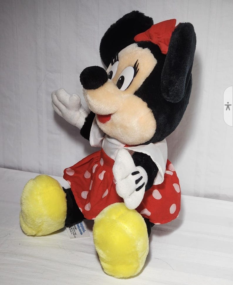 May include: A plush Minnie Mouse doll, featuring black fur, a red polka-dot skirt, and a matching bow. The doll has large yellow feet, white gloves, and a white collar. The doll is sitting and appears to be waving.