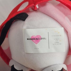 Pu&ograve; includere: Un peluche con corpo bianco, orecchie nere e un fiocco rosso a pois. L'etichetta recita "KOHL'S cares" con un logo a forma di cuore e "&copy;Disney". Il giocattolo ha dettagli in bianco e nero.