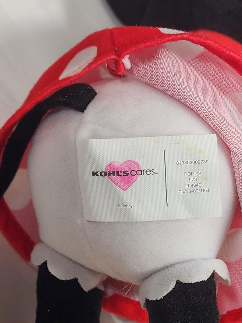 Pu&ograve; includere: Un peluche con corpo bianco, orecchie nere e un fiocco rosso a pois. L'etichetta recita "KOHL'S cares" con un logo a forma di cuore e "&copy;Disney". Il giocattolo ha dettagli in bianco e nero.