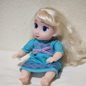 Puede incluir: Mu&ntilde;eca con cabello rubio y ojos azules, vestida con un vestido turquesa con un estampado floral morado y un cintur&oacute;n de terciopelo morado. La mu&ntilde;eca est&aacute; sentada con los brazos y las piernas extendidos.