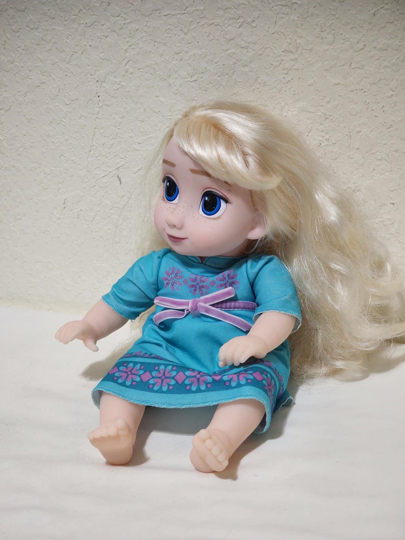 Puede incluir: Mu&ntilde;eca con cabello rubio y ojos azules, vestida con un vestido turquesa con un estampado floral morado y un cintur&oacute;n de terciopelo morado. La mu&ntilde;eca est&aacute; sentada con los brazos y las piernas extendidos.
