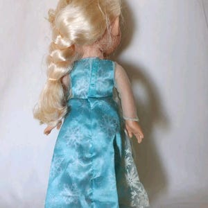 Op de afbeelding: Een blonde pop die een blauwe jurk draagt met een sneeuwvlokkenpatroon. De jurk heeft een doorschijnende overlay met sneeuwvlokken. De pop heeft een gevlochten paardenstaart.
