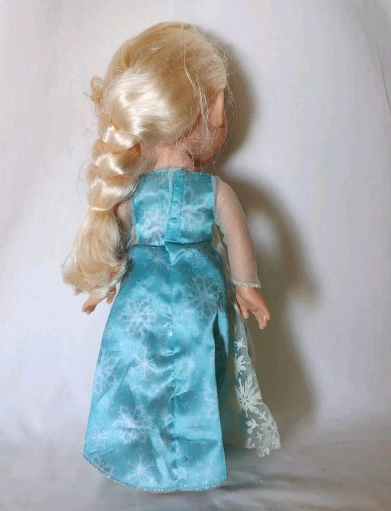 Op de afbeelding: Een blonde pop die een blauwe jurk draagt met een sneeuwvlokkenpatroon. De jurk heeft een doorschijnende overlay met sneeuwvlokken. De pop heeft een gevlochten paardenstaart.