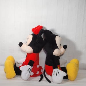 Pu&ograve; includere: Due bambole di peluche dei personaggi Disney, Topolino e Minnie, sono sedute schiena contro schiena. Minnie indossa un vestito rosso a pois bianchi e un fiocco rosso. Topolino indossa pantaloncini rossi. Entrambi hanno guanti bianchi e scarpe gialle.