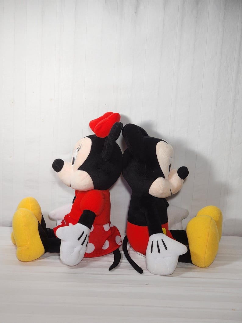 Pu&ograve; includere: Due bambole di peluche dei personaggi Disney, Topolino e Minnie, sono sedute schiena contro schiena. Minnie indossa un vestito rosso a pois bianchi e un fiocco rosso. Topolino indossa pantaloncini rossi. Entrambi hanno guanti bianchi e scarpe gialle.