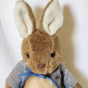May include: A brown plush Peter Rabbit toy wearing a blue jacket with a white collar and a blue bow tie. The toy has brown fur, long ears, and black eyes.