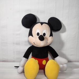 Pu&ograve; includere: Un pupazzo di peluche di Topolino, con orecchie nere, corpo nero e viso e mani bianche. La bambola ha pantaloncini rossi e scarpe gialle. Il giocattolo &egrave; seduto in posizione eretta su uno sfondo bianco.