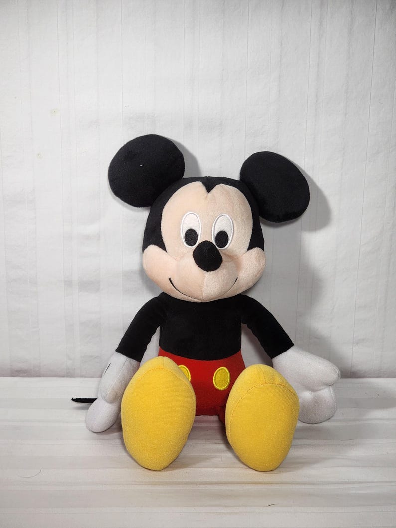 Pu&ograve; includere: Un pupazzo di peluche di Topolino, con orecchie nere, corpo nero e viso e mani bianche. La bambola ha pantaloncini rossi e scarpe gialle. Il giocattolo &egrave; seduto in posizione eretta su uno sfondo bianco.