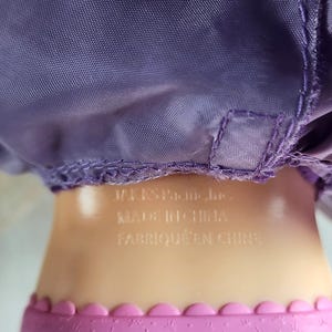 May include: Close-up of a doll's body. The doll has a purple satin dress and a pink skirt with a scalloped edge. The text "JAKKS Pacific Inc. MADE IN CHINA FABRIQU&Eacute; EN CHINE" is printed on the doll's body.