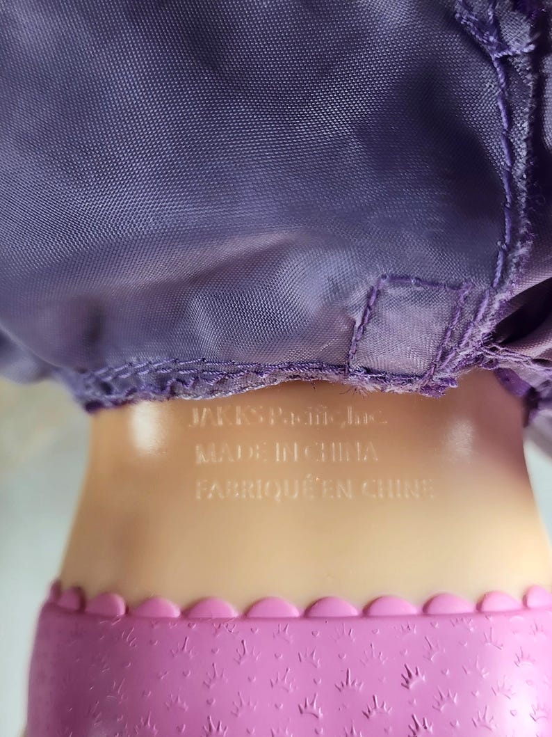 May include: Close-up of a doll's body. The doll has a purple satin dress and a pink skirt with a scalloped edge. The text "JAKKS Pacific Inc. MADE IN CHINA FABRIQU&Eacute; EN CHINE" is printed on the doll's body.