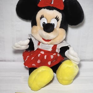 May include: A plush Minnie Mouse doll with black ears, a red bow, and a red polka-dot dress. The doll has white gloves and yellow feet. A measuring tape is at the bottom of the image.