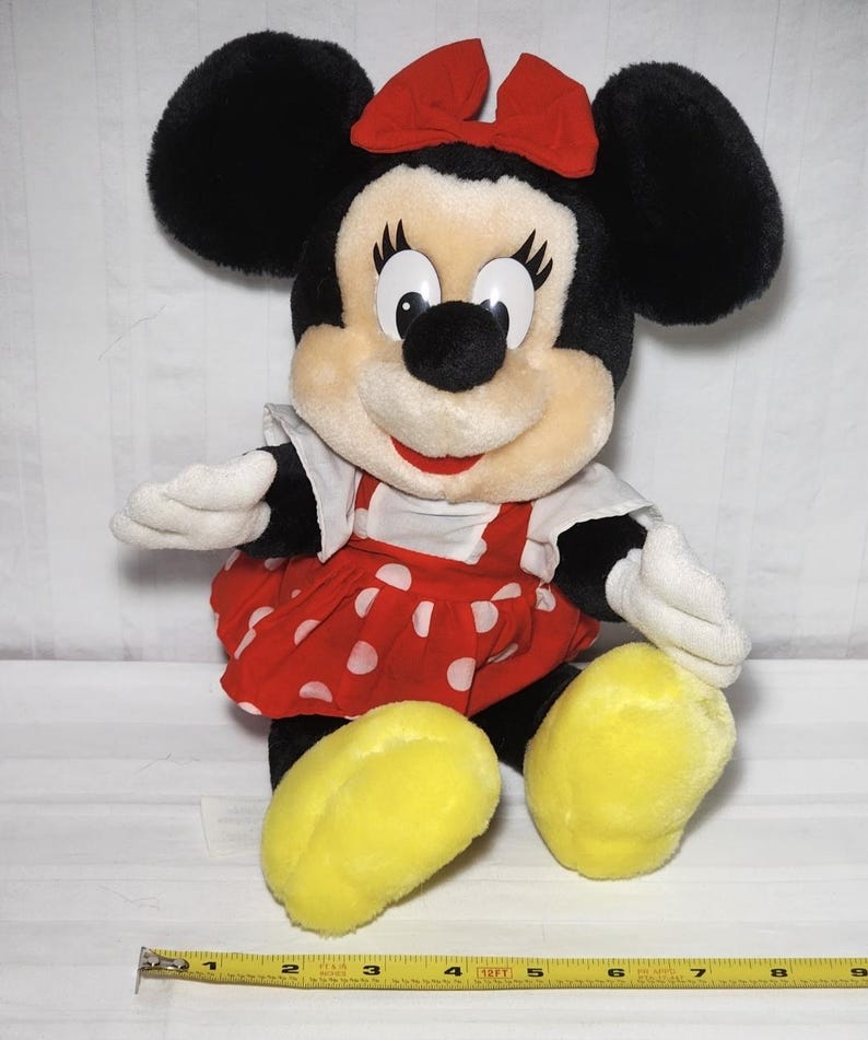May include: A plush Minnie Mouse doll with black ears, a red bow, and a red polka-dot dress. The doll has white gloves and yellow feet. A measuring tape is at the bottom of the image.