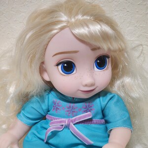 Puede incluir: Mu&ntilde;eca con cabello rubio y ojos azules. La mu&ntilde;eca viste un vestido turquesa con un lazo morado y detalles florales. La cara de la mu&ntilde;eca tiene pecas y una suave sonrisa.