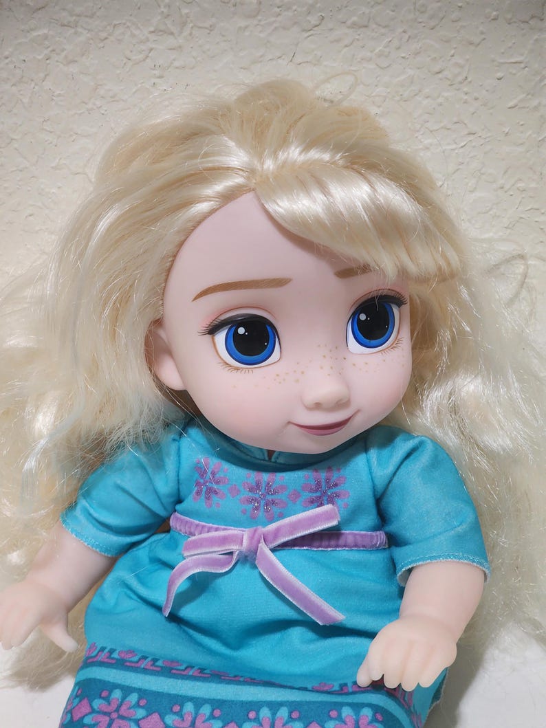 Puede incluir: Mu&ntilde;eca con cabello rubio y ojos azules. La mu&ntilde;eca viste un vestido turquesa con un lazo morado y detalles florales. La cara de la mu&ntilde;eca tiene pecas y una suave sonrisa.