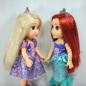 May include: Two dolls of Disney princesses, one with long blonde hair and a purple dress, the other with red hair and a mermaid-style outfit. Both dolls wear tiaras and purple shoes, holding hands. The dolls are standing in front of a white background.