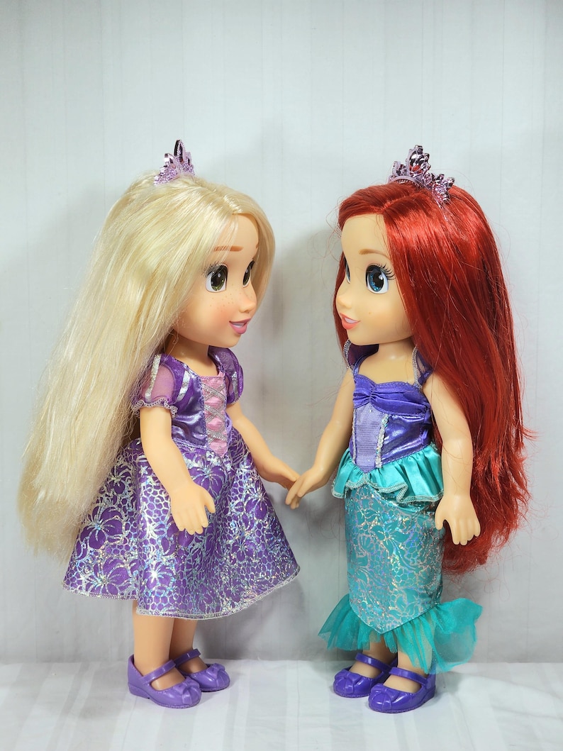 May include: Two dolls of Disney princesses, one with long blonde hair and a purple dress, the other with red hair and a mermaid-style outfit. Both dolls wear tiaras and purple shoes, holding hands. The dolls are standing in front of a white background.