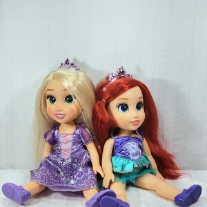 May include: Two Disney princess dolls, Rapunzel and Ariel, sit side-by-side. Rapunzel has long blonde hair and wears a purple dress and shoes. Ariel has red hair, a purple top, and a teal skirt. Both dolls wear tiaras.
