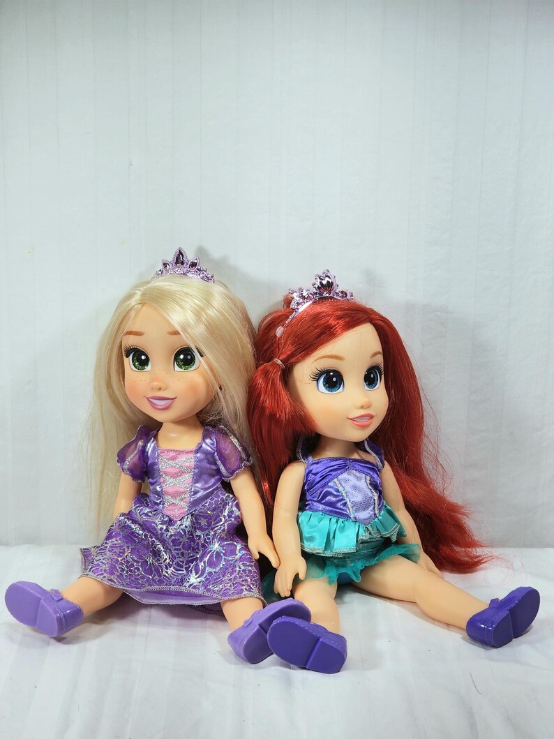 May include: Two Disney princess dolls, Rapunzel and Ariel, sit side-by-side. Rapunzel has long blonde hair and wears a purple dress and shoes. Ariel has red hair, a purple top, and a teal skirt. Both dolls wear tiaras.