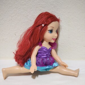 Puede incluir: Mu&ntilde;eca de un personaje con largo cabello rojo, ojos azules y un vestido morado y azul. La mu&ntilde;eca est&aacute; sentada con las piernas separadas. El cabello de la mu&ntilde;eca est&aacute; peinado con una peque&ntilde;a trenza.