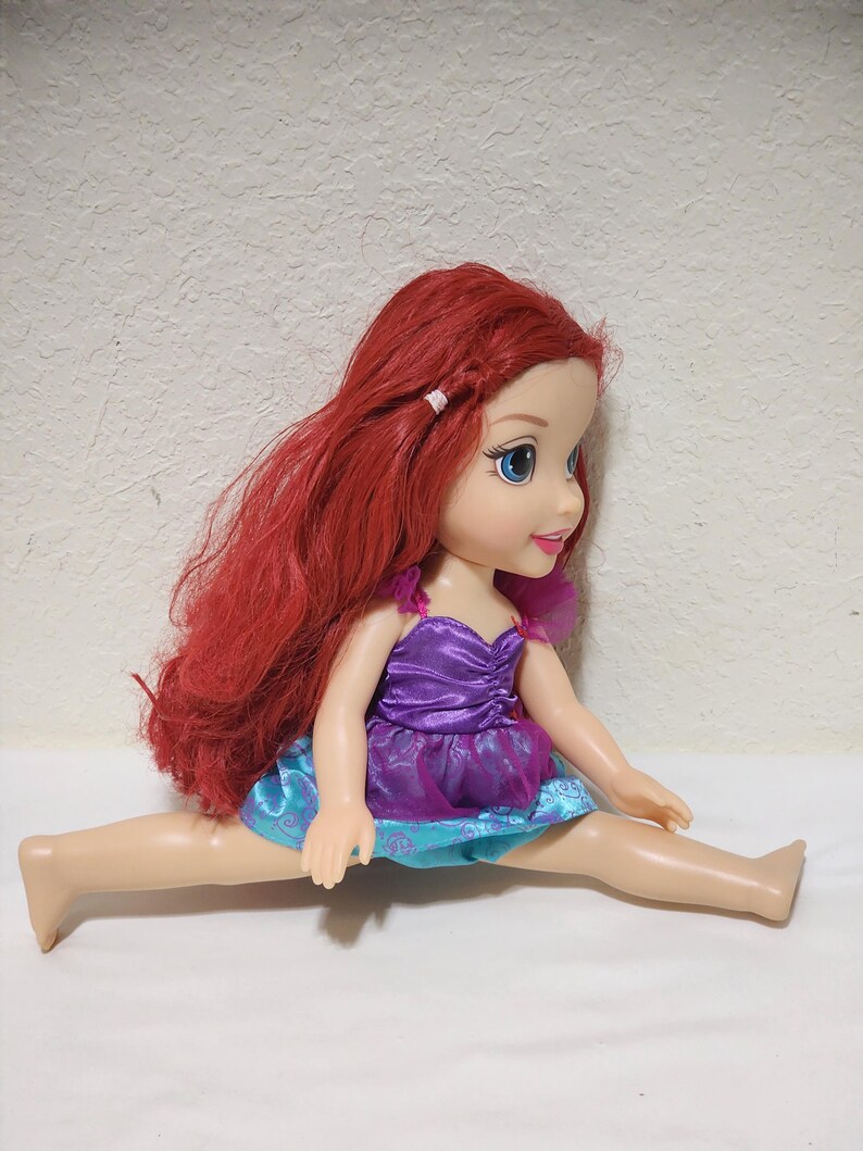 Puede incluir: Mu&ntilde;eca de un personaje con largo cabello rojo, ojos azules y un vestido morado y azul. La mu&ntilde;eca est&aacute; sentada con las piernas separadas. El cabello de la mu&ntilde;eca est&aacute; peinado con una peque&ntilde;a trenza.