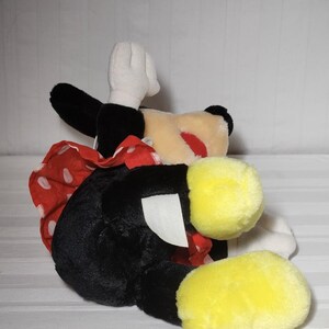 May include: A plush Minnie Mouse doll, featuring a black body, white gloves, and yellow feet. The doll has a red and white polka dot bow and a cream-colored face with a red nose. The doll is lying on its side.