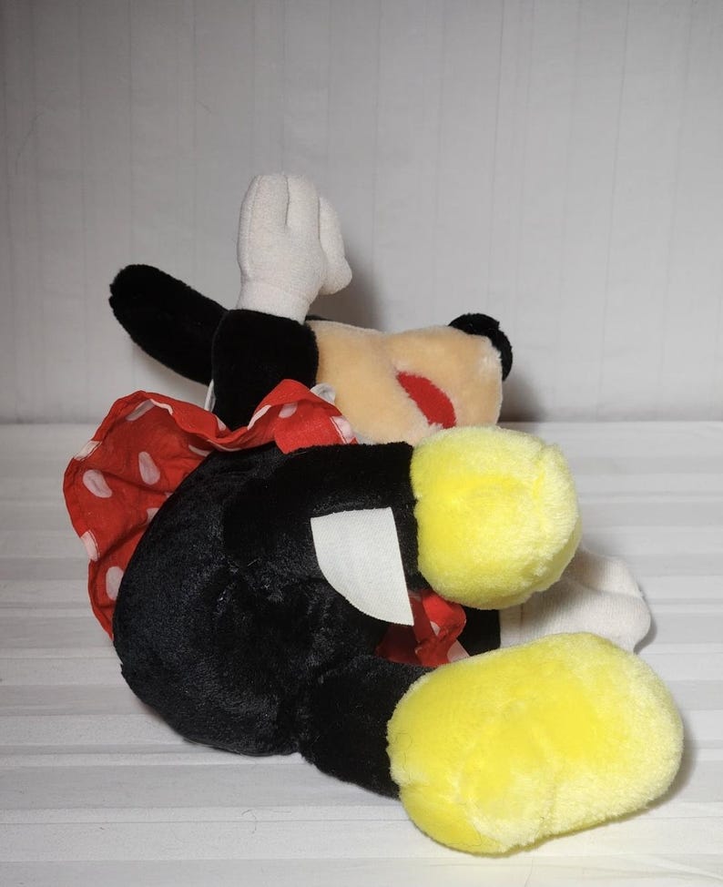 May include: A plush Minnie Mouse doll, featuring a black body, white gloves, and yellow feet. The doll has a red and white polka dot bow and a cream-colored face with a red nose. The doll is lying on its side.