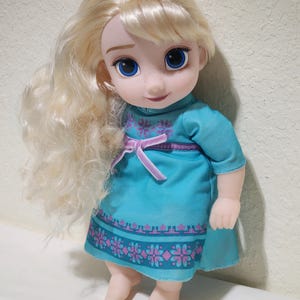 Puede incluir: Mu&ntilde;eca de un personaje con cabello rubio y ojos azules, vestida con un vestido turquesa con un estampado floral rosa y morado. La mu&ntilde;eca tiene una expresi&oacute;n sonriente y est&aacute; de pie.