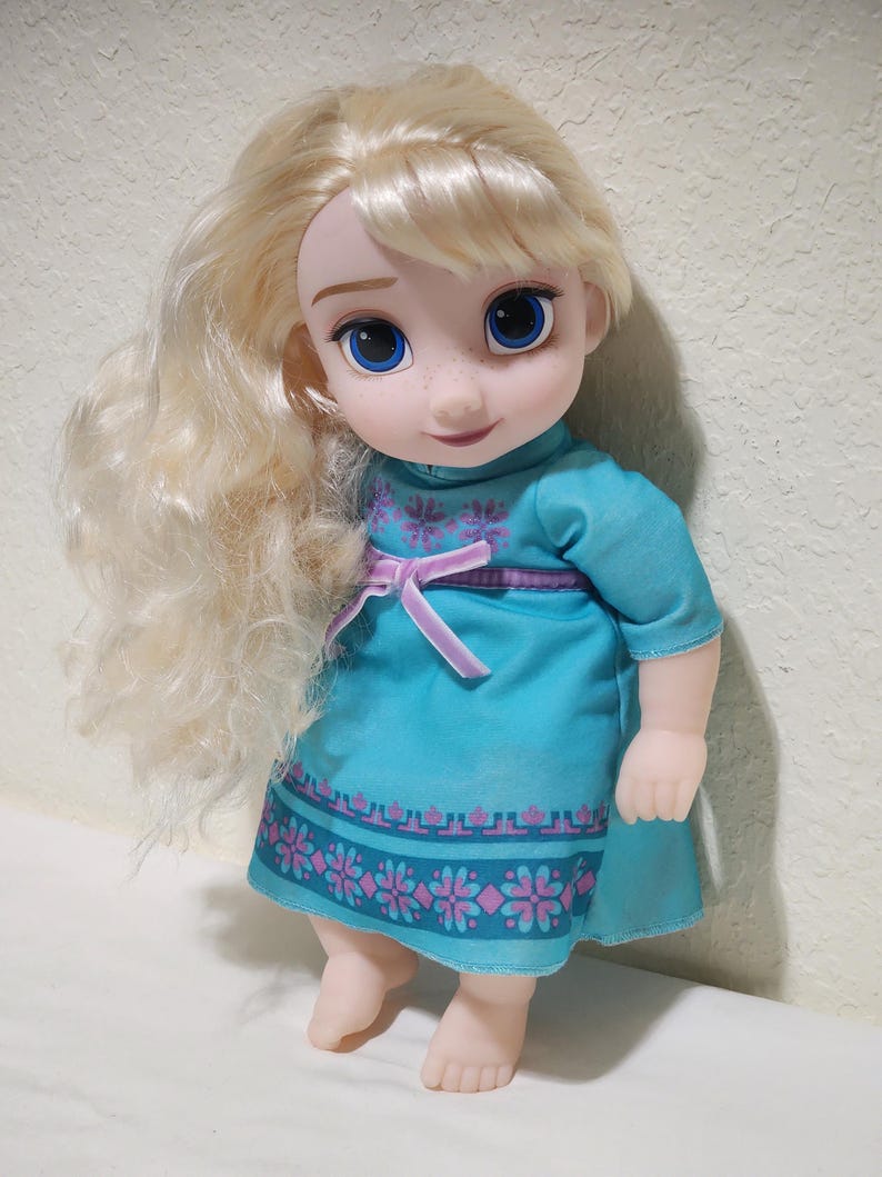 Puede incluir: Mu&ntilde;eca de un personaje con cabello rubio y ojos azules, vestida con un vestido turquesa con un estampado floral rosa y morado. La mu&ntilde;eca tiene una expresi&oacute;n sonriente y est&aacute; de pie.
