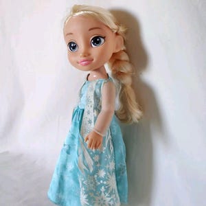 Op de afbeelding: Een blonde pop die een lichtblauwe jurk draagt met een wit sneeuwvlokkenpatroon. De pop heeft een gevlochten kapsel en blauwe ogen.
