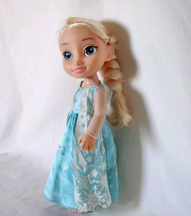 Op de afbeelding: Een blonde pop die een lichtblauwe jurk draagt met een wit sneeuwvlokkenpatroon. De pop heeft een gevlochten kapsel en blauwe ogen.