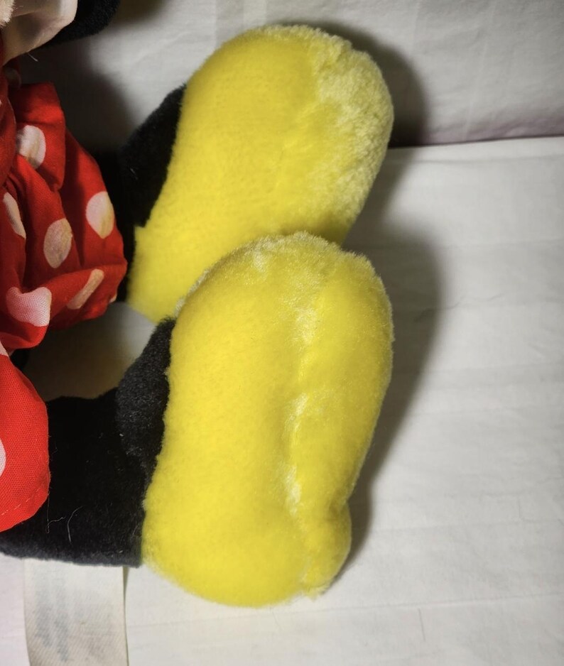 May include: Close-up of a plush toy's yellow feet and black legs. The feet are a bright yellow color, and the legs are black. A red polka-dot dress is visible on the left side of the image. The toy is likely a character from a popular cartoon.