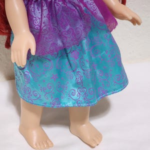 Puede incluir: Una mu&ntilde;eca con un vestido morado y turquesa con un dise&ntilde;o estampado. La mu&ntilde;eca tiene el pelo rojo y est&aacute; de pie sobre una superficie blanca. El vestido tiene un aspecto brillante y satinado.