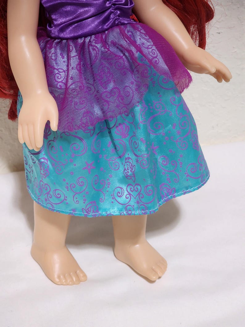 Puede incluir: Una mu&ntilde;eca con un vestido morado y turquesa con un dise&ntilde;o estampado. La mu&ntilde;eca tiene el pelo rojo y est&aacute; de pie sobre una superficie blanca. El vestido tiene un aspecto brillante y satinado.
