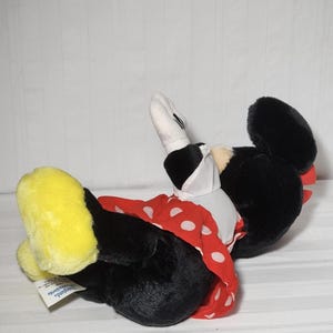 May include: A plush Minnie Mouse doll, lying on its side. The doll features a black body, large black ears, and a red dress with white polka dots. Yellow shoes and white gloves complete the look. The doll is a classic Disney character.