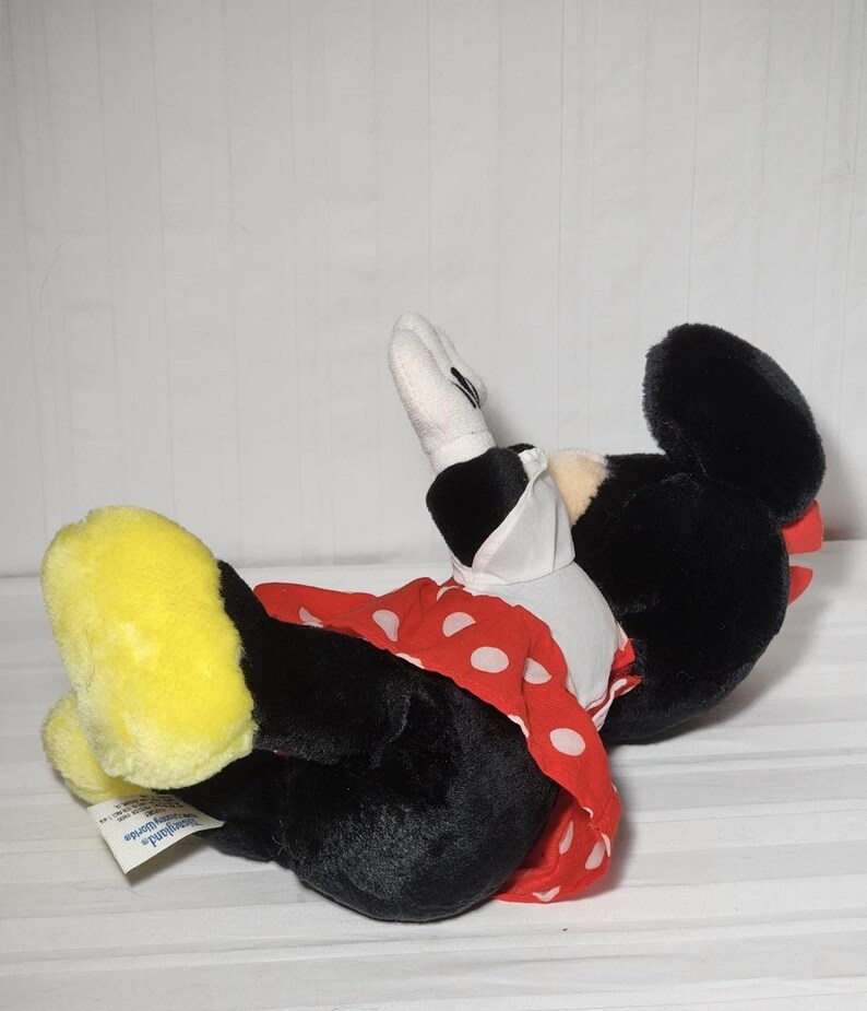 May include: A plush Minnie Mouse doll, lying on its side. The doll features a black body, large black ears, and a red dress with white polka dots. Yellow shoes and white gloves complete the look. The doll is a classic Disney character.