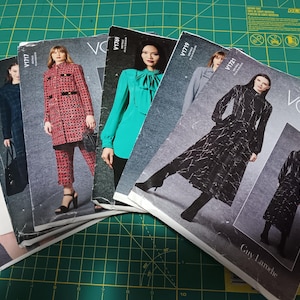 May include: A collection of Vogue Patterns, each displaying a different fashion design. The covers feature women modelling outfits such as a red and black patterned coat and trousers, a teal blouse, and a black and white patterned dress. The patterns are on a green cutting mat.