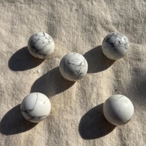 Può includere: Cinque pietre di howlite bianche, lisce e sferiche, disposte in un motivo pentagonale. Ogni pietra presenta un distinto motivo naturale di venature grigie, che crea un aspetto marmorizzato sulle superfici levigate e arrotondate.