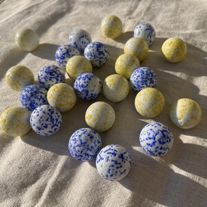 Op de afbeelding: Een verzameling van 20 glazen knikkers met een witte basis en blauwe en gele stippen. De knikkers zijn gerangschikt op een wit stoffen oppervlak.