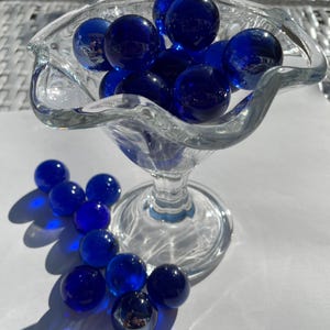 Op de afbeelding: Een glazen kom gevuld met blauwe knikkers, met meer blauwe knikkers verspreid over een wit oppervlak.