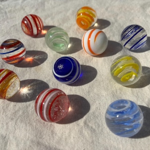 Op de afbeelding: Een verzameling van tien bolvormige glazen knikkers met diverse ontwerpen, waaronder helder glas met interne wervelingen van rood, blauw, oranje, geel en wit, die in elk afzonderlijk bolvormig speelgoed verschillende patronen creëren.