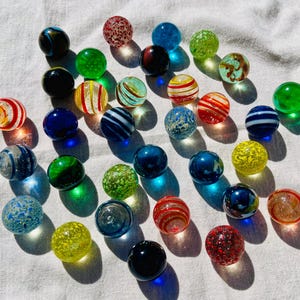 May include: A collection of colourful glass marbles in various patterns and colours, including red, blue, green, yellow, and white. The marbles are arranged on a white fabric surface.
