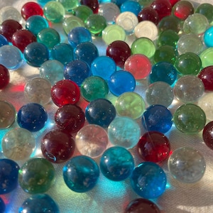 Op de afbeelding: Een verzameling kleurrijke glazen knikkers in tinten blauw, groen, rood en helder. De knikkers zijn verspreid over een wit oppervlak.