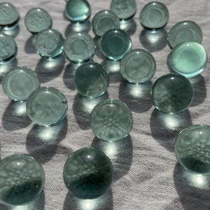 Può includere: Una collezione di biglie di vetro blu turchese sparse su una superficie di tessuto bianco. Le biglie sono rotonde e hanno una finitura liscia e lucida.
