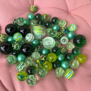 Op de afbeelding: Een verzameling diverse glazen knikkers in verschillende tinten groen, zwart en helder glas. De knikkers hebben uiteenlopende patronen, waaronder wervelingen, effen kleuren en gespikkelde ontwerpen, sommige met interne gele en blauwe strepen.
