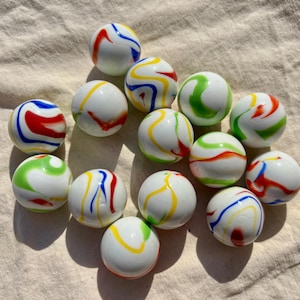 Op de afbeelding: Een verzameling van dertien bolvormige glazen knikkers, elk met een gladde witte ondoorzichtige basis met wervelende, veelkleurige patronen in tinten rood, blauw, groen en geel, gerangschikt in een losse groep op een textieloppervlak.