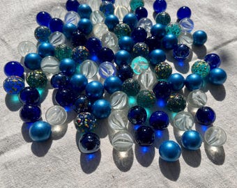 May include: A collection of blue, white and green glass marbles. The marbles are scattered on a white fabric surface.
